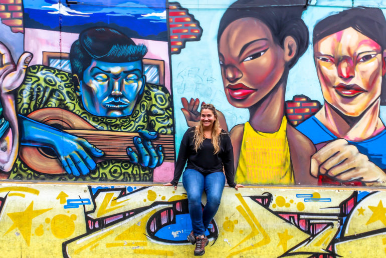 Girl sitting on a half wall in front of a mural. 